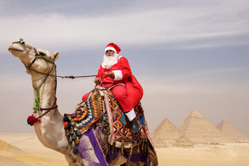 Naklejka premium Camel rider in full red Santa costume holding reins atop a colorfully saddled dromedary camel with the iconic Giza pyramids rising behind in the sandy