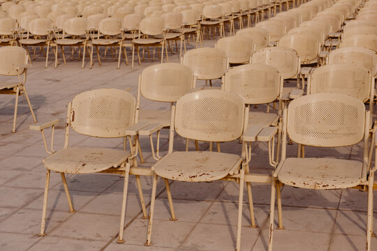 Rows of empty weathered cream metal chairs with rust detail arranged on a tiled plaza at an open air outdoor venue before a public event or concert