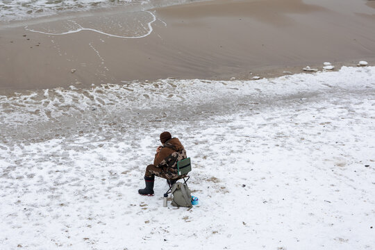 Fisherman sitting alone on snow covered beach by sea on overcast winter day