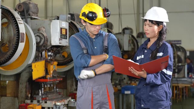 Industrial worker and female engineer reviewing red folder and manual beside heavy machinery and machinery in factory workplace manufacturing setting