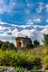 Obraz premium Ancient roman archway ruins set in lush mediterranean landscape. Roman arch standing amidst lush Mediterranean vegetation and wild greenery under dramatic blue sky with fluffy white clouds.