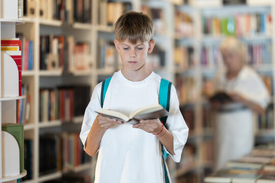 Interested teen male visitor holding a book in public library with large stock
