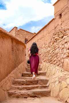 Woman climbing rustic stone steps between earthen kasbah walls at Ait Benhaddou exploring historic fortified village architecture and desert heritage under a clear blue sky