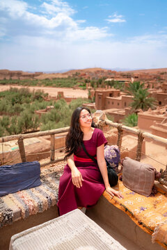 Young Latin woman relaxing on a rooftop terrace enjoying panoramic views of the historic Ait Benhaddou kasbah and surrounding Moroccan landscape during her travel adventure
