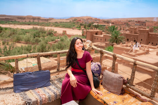Young woman traveler enjoying the panoramic view of the ancient Ait Benhaddou ksar in Morocco sitting comfortably on a traditional rooftop terrace on a sunny day