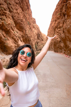 Young smiling Latin woman taking a happy selfie standing on a road in Todra Gorge Morocco celebrating a thrilling travel adventure surrounded by towering orange canyon walls