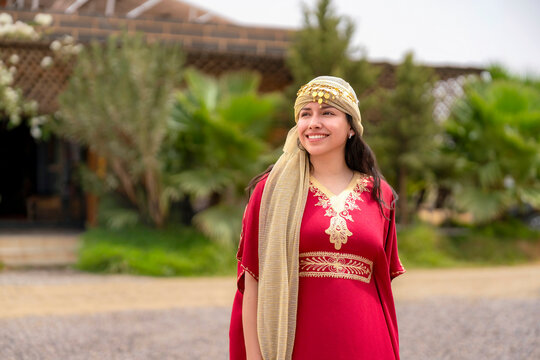 Young smiling woman wearing a traditional red kaftan with gold embroidery and a decorative turban looking away while enjoying a cultural experience in Marrakech Morocco