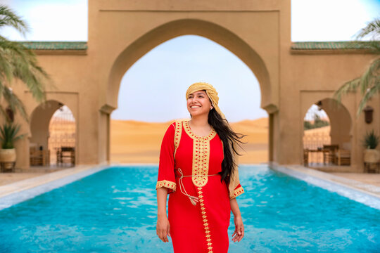 Young smiling woman wearing traditional red caftan and headscarf standing by a refreshing swimming pool at an exotic desert resort with traditional architecture and golden dunes in the background