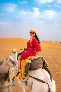 Young smiling woman enjoying camel riding during a desert safari adventure wearing traditional red dress and head wrap in the vibrant dunes under a blue sky