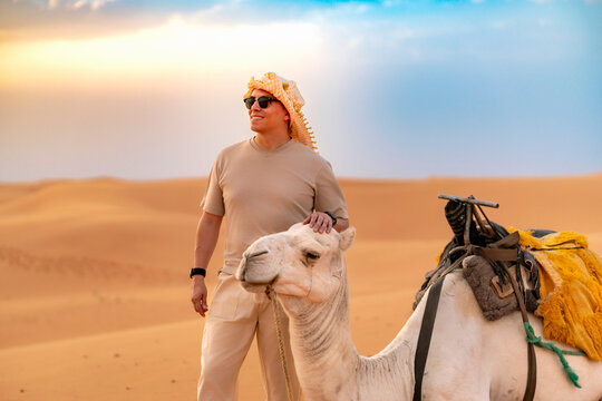 Smiling man in traditional keffiyeh and sunglasses gently pets a white camel amid golden Sahara dunes at sunrise in Morocco enjoying an exotic desert adventure and peaceful moment