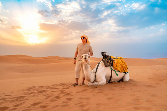 Young man wearing a keffiyeh and sunglasses standing next to a resting camel on a sand dune experiencing a Moroccan desert adventure during golden hour