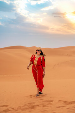 Young smiling woman wearing a red Moroccan kaftan and head covering walking through sandy desert dunes with a beautiful sunset sky embracing travel and cultural experience