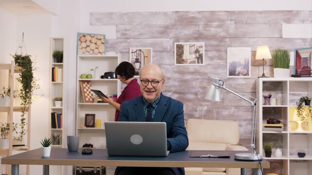 Smiling older man reacts to good news on his laptop while a woman uses a tablet behind him, combining optimism, online engagement and active aging.