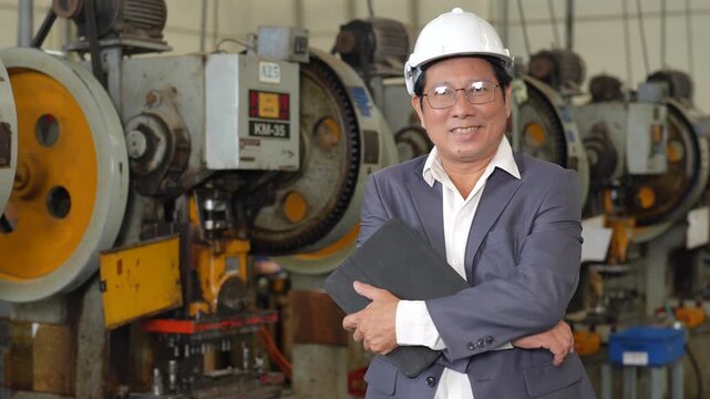 Asian adult man engineer wearing safety helmet beside metal press machinery industrial factory, manufacturing worker conducting safety inspection with protective eyewear, folder, confidence