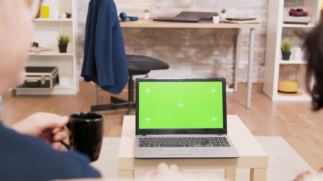 Seen from behind, a senior couple sit together and wave toward a laptop with a chroma key display, creating a home video call and screen replacement concept.
