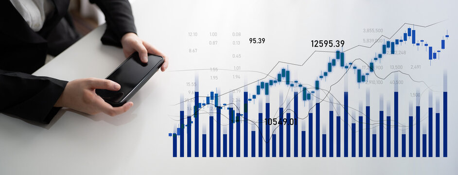 A business professional holds a smartphone while analyzing financial graphs and market trends on a table surface, representing modern finance analytics. Scalp