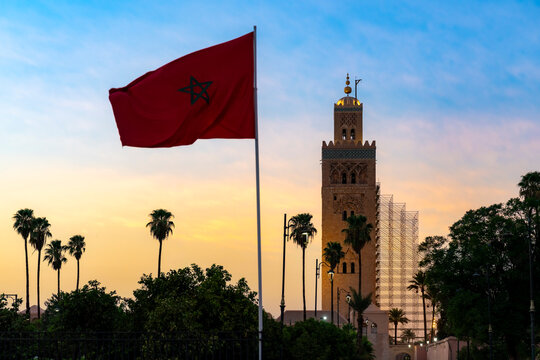 Moroccan flag flying prominently over the Koutoubia Mosque minaret a historic landmark in Marrakech during a colorful sunset surrounded by green palm trees