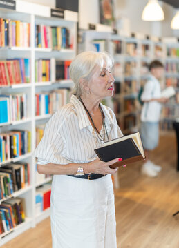Focused mature lady browsing through literature volumes on shelves in cozy bookshop