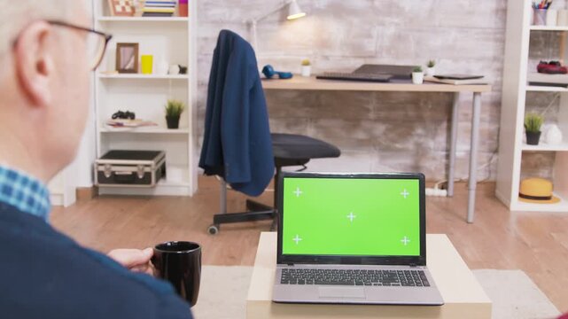 Seen from behind, a senior couple sit on a sofa as the man waves toward a laptop with a chroma key display, combining a home video call with screen replacement, compositing and online communication.