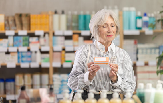 Mature woman stands in front of the shelves in the pharmacy and chooses paracetamol from different manufacturers. Pensioner in a European pharmacy does shopping