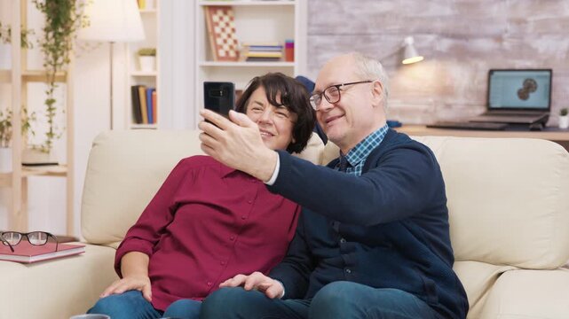 Smiling older couple lean together as the man extends a smartphone for a selfie, sharing a cheerful moment of connection and digital leisure at home.