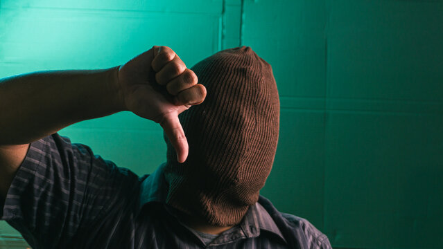 Faceless Indonesian adult male wearing a reversed brown balaclava displaying a prominent thumbs down gesture to signify strong disapproval or failure. The completely anonymous Asian