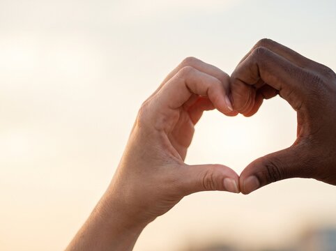 Two diverse hands, one light-skinned and one dark-skinned, forming a heart shape against a soft, bright background, symbolizing love and unity.
