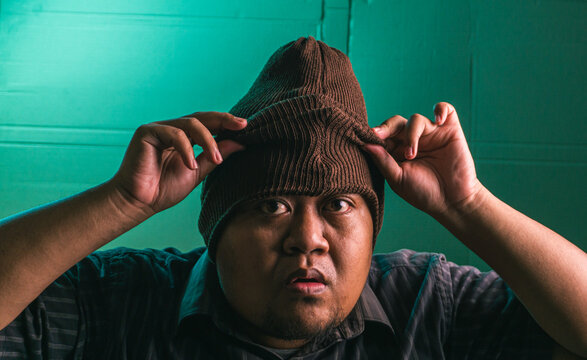 Serious Indonesian adult male pulling a brown knit beanie firmly down over the forehead using both hands while staring intensely directly into the camera lens. The intimidating Asian