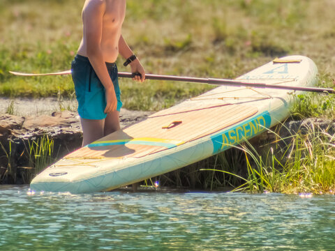 Canmore, Alberta, Canada. September 9, 2026. A young boy standing on the edge of a grassy lake shore holding an Ascend paddleboard.