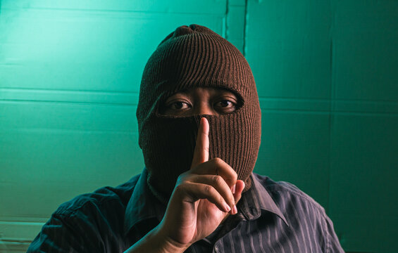 Secretive Indonesian adult male wearing brown balaclava mask making silence gesture with index finger over mouth, mysterious criminal person isolated against dark green studio wall