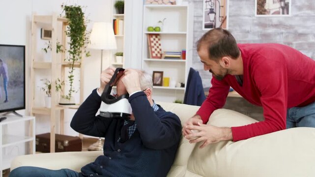 Younger man helps a senior learn to use a VR headset at home, creating a hands-on lesson about digital learning, technology assistance, active aging and supportive intergenerational guidance.
