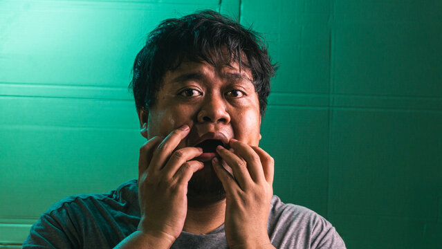 An astonished young indonesian male touches the mouth area with the fingers, displaying a wide open jaw and immense disbelief against a green textured background. The subject stares