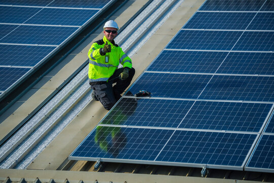 A photovoltaic technician wearing a safety vest and hard hat precisely crouches over the panels for a final evening test on a commercial factory rooftop photovoltaic construction site.
