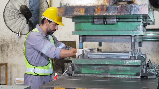 Industrial worker wearing safety helmet and reflective vest operating metal forming machine and industrial press inside factory, focused metal fabrication task beside heavy machinery and protective