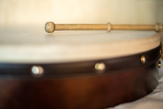 The two-sided stick, tipper, or beater lies on the white goat-skin-covered hand drum. The drum's frame is dark brown. There's a black edging with silver pins keeping the skin taut. The hand drum is a 