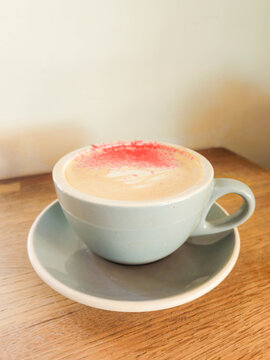 A light blue colored cup and saucer filled with hot peppermint mocha coffee sit on a wooden table. The drink has red peppermint crystals on top of its froth, with a leaf pattern in the milk foam. 