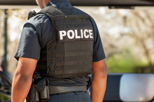A male police officer wears a taser and a handgun. The law enforcement officer wears a black bulletproof vest, a black shirt, and dark pants. The word police is on his back. 