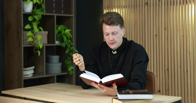 Priest in cassock with rosary reading Holy Bible at wooden table indoors