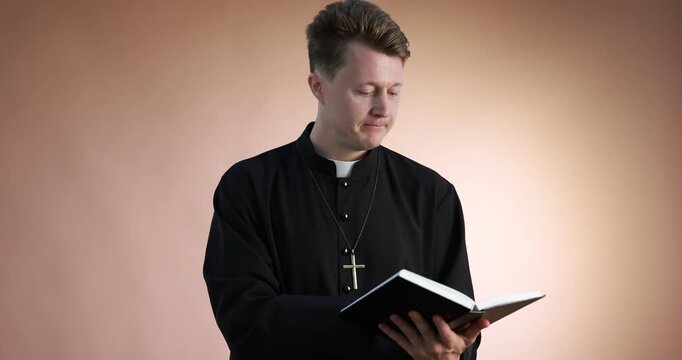 Priest in cassock reading Holy Bible on pale brown background