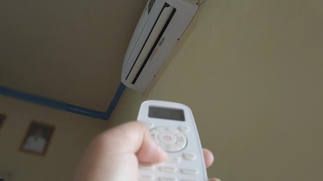A person using an air conditioner remote control to turn on an indoor air conditioning unit. The scene represents home comfort, temperature control, cooling technology, and everyday household activiti