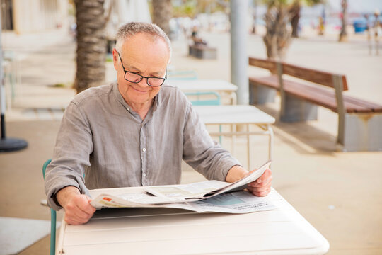 An elderly man in glasses reads an unfolded newspaper
