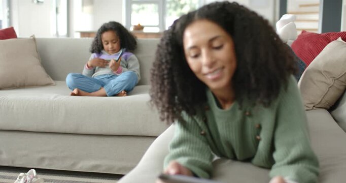African mother tapping tablet while camera shifting focus, daughter on sofa scrolling smartphone