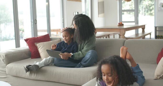 African American mother with two child daughters positioning tablet, guiding child learning on sofa