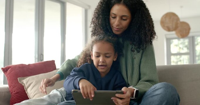 African American mom and child on sofa using tablet, mom tilting it guiding child tapping bonding