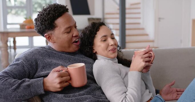 African American couple on grey sofa at home with pink mugs, man telling joke, woman laughing