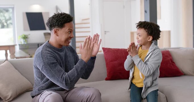 African American man raising palms, boy mirroring clapping and high-fives on sofa for bonding