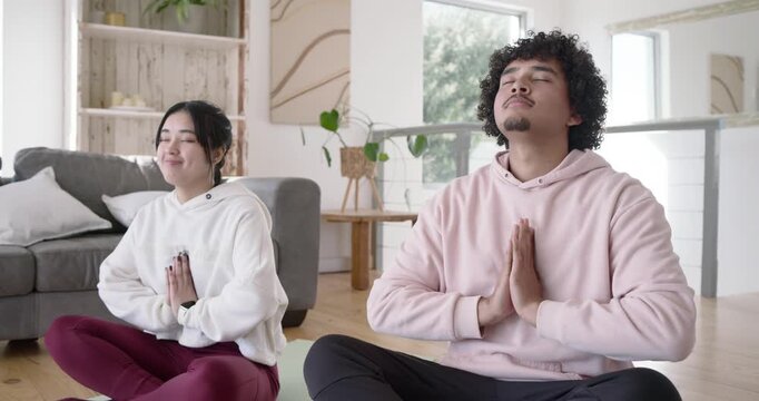 Diverse couple smiling, sitting on mats in living room, pressing palms, meditating, high-fiving