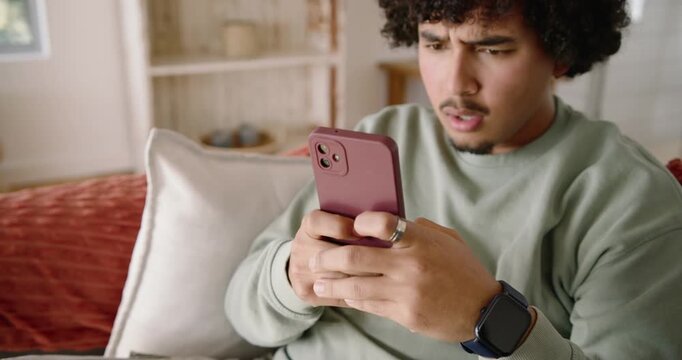 Adult man sitting on couch at home, reading mauve smartphone message and reacting, tapping thumbs