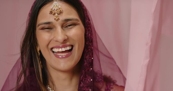 Indian woman smiling, laughing, settling into soft smile facing camera in studio with maroon veil