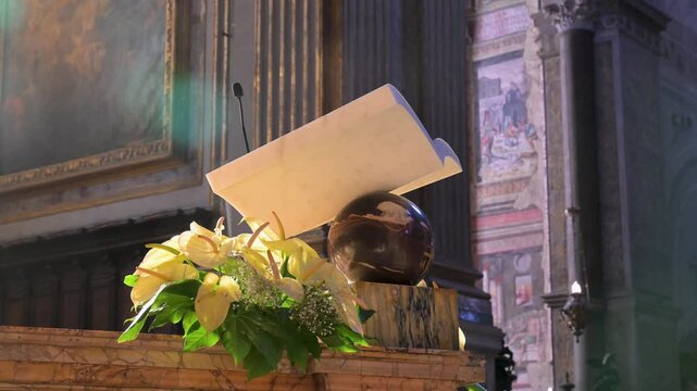 Marble lectern with open book, microphone, and yellow lilies catches warm sunlight inside an ornate cathedral in Naples.